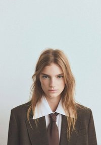 Blazer oversize marrone a quadri con ampio colletto bianco, indossato sopra una camicia bianca e una cravatta marrone. La modella ha capelli lunghi e mossi.