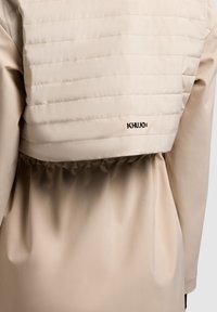 Bež prešita jakna od zadaj z elastičnim pasom in majhnim črnim logotipom »Khujo« na sredini zgornjega dela hrbta.