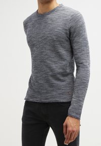 Gråblå tröja med en texturerad stickning, rund halsringning och fransad fåll. Har lång ärm och en liten märke nära botten.