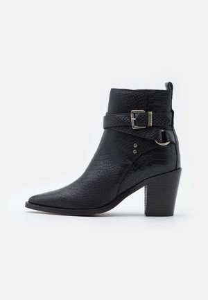Bottine noire texturée avec bout pointu, talon bloc, sangle décorative et boucle argentée sur fond blanc.