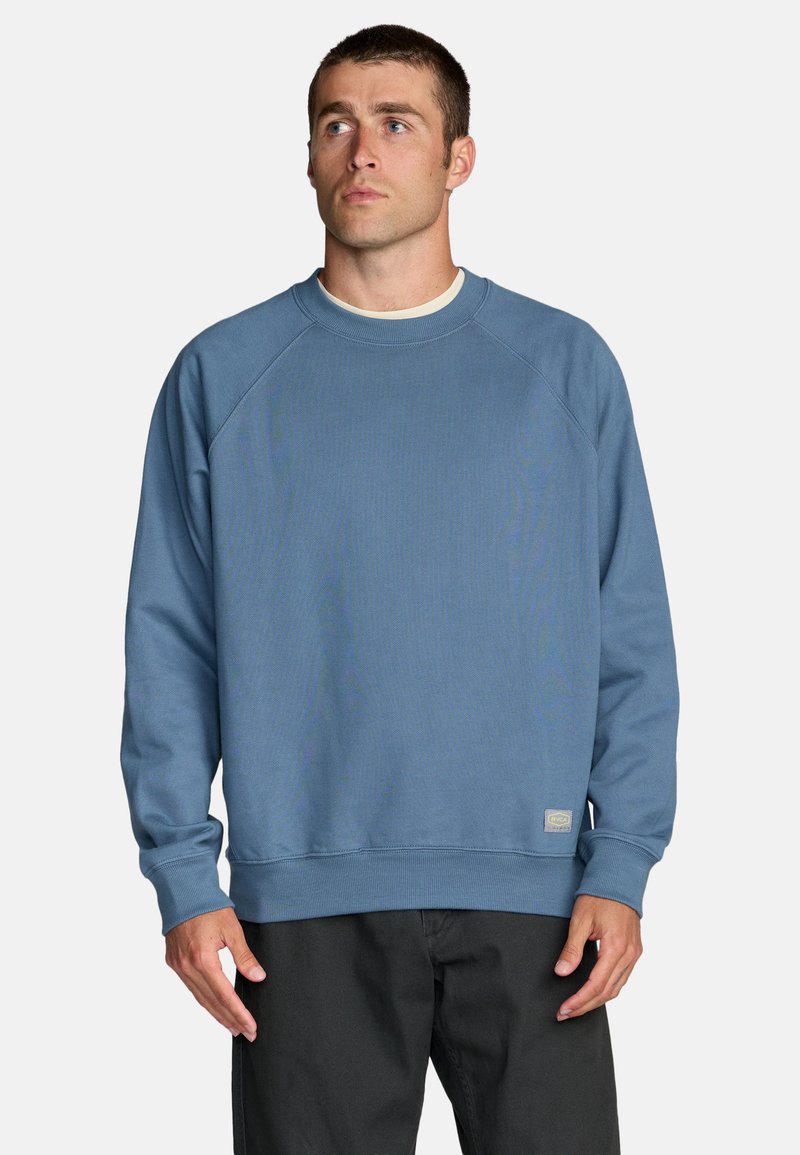 Mężczyzna ubrany w niebieską bluzę z długim rękawem i czarne spodnie, lekko spoglądający w prawo na jednolitym białym tle.