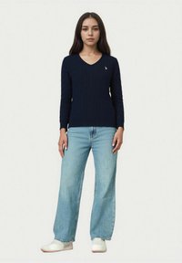 Pulover bleumarin tricotat cu guler în V, având un model cu torsade. Asortat cu blugi largi, de culoare albastru deschis, și pantofi albi.