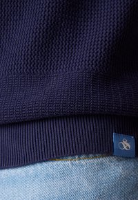 Mörkblå stickad tröja med texturerat mönster och ribbad fåll, med en liten blå märkesetikett synlig på sidan.
