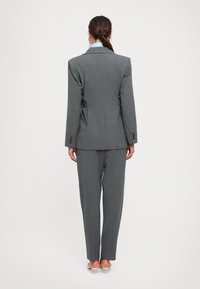 Woman with dark hair tied back wearing a gray tailored suit and light blue shirt, standing facing away, on a plain white background.