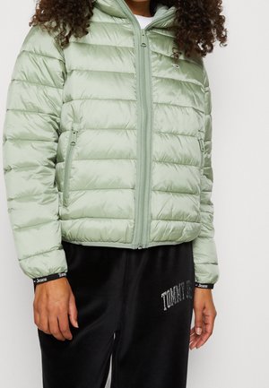Personne portant une veste matelassée vert clair avec des poches zippées et un pantalon noir avec le texte « Tommy Jeans » sur le poignet et la cuisse.
