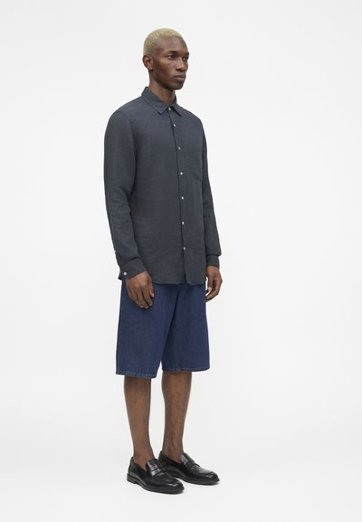 Μαύρος αρσενικός μοντέλο με κοντά ξανθά μαλλιά, φοράει σκούρο πουκάμισο με κουμπιά, μπλε σορτς μέχρι το γόνατο και μαύρα μοκασίνια, στέκεται μπροστά από άσπρο φόντο.