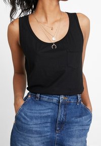 Svart ärmlös tanktopp med rund halsringning och ett litet bröstfack, tillsammans med högmidjade blå denimjeans.