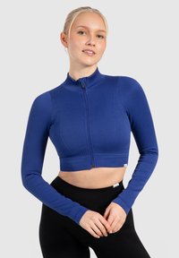 Geribbelde, cropped lange mouw jack in koningsblauw met een hoge kraag en een voorkant met rits. Heeft een getailleerd ontwerp en een gladde textuur. Zwarte broek eronder.