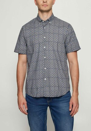 Mand iført en kortærmet skjorte med knapper med et cirkulært mønster i marineblåt, hvidt og beige, kombineret med blå jeans.