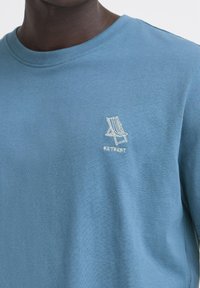Lyseblå bomulds T-shirt med et lille hvidt broderet ikon af en liggestol og ordet "RETREAT" under. Rund hals design.