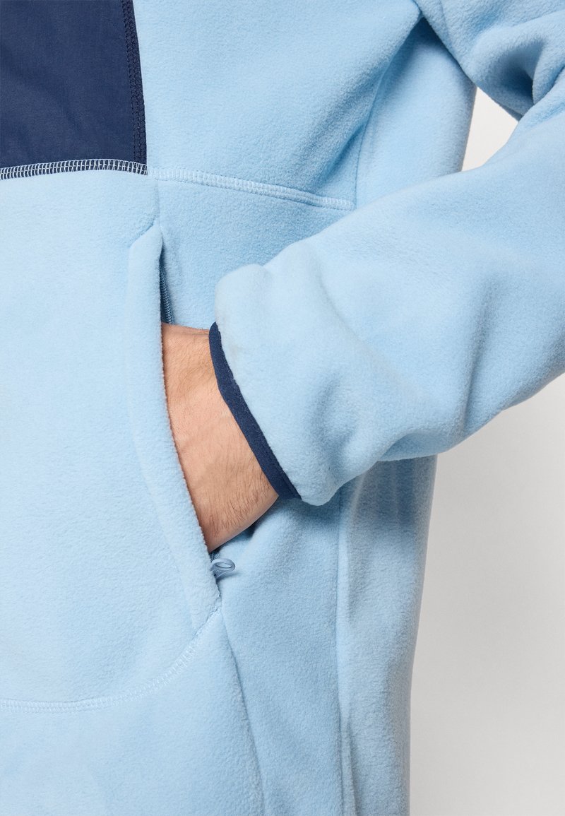 Jasnoniebieska polarowa kurtka z granatowym akcentem. Posiada przednią kieszeń, elastyczne mankiety oraz miękką teksturę.