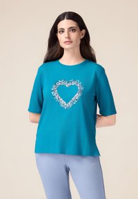 Camiseta de manga corta en color azul verdoso con un diseño de corazón hecho de brillantes lentejuelas azules. Corte casual con escote redondo.