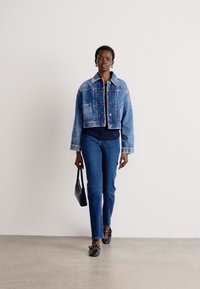 Giacca di jeans tagliata con lavaggio blu, due tasche frontali, cuciture bianche. Abbinata a jeans scuri e scarpe basse nere, mentre tiene una borsa nera.