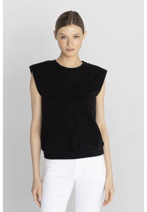 Kobieta nosząca czarny top bez rękawów z wyściełanymi ramionami i białe spodnie, stojąca na jednolitym jasnoszarym tle.