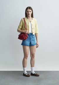 Noor naine kollast lühikeste varrukatega jopet, valget särki, siniseid teksapükse, valgeid sukki ja leopardimustriga kingi kandmas, punane käekott õlal.