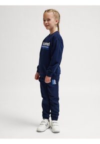 Completo in felpa blu navy e pantaloni realizzato in tessuto morbido, con stampa del logo. Abbinato a scarpe da ginnastica bianche, con trecce laterali visibili.