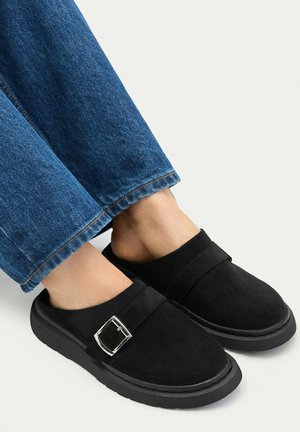 Füße in schwarzen Schlupfschuhen mit silberner Schnallendetail und dunkelblauer Jeans vor hellem, schlichten Hintergrund.