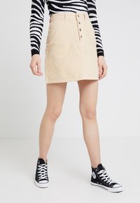 Minifalda de denim beige claro con botones delanteros y bolsillos, combinada con zapatillas negras de caña alta, sobre un fondo liso.