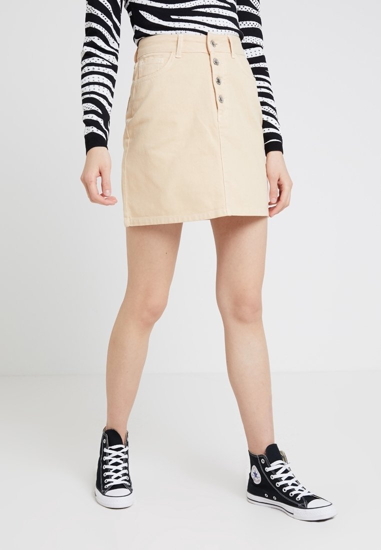 Minifalda de denim beige claro con botones delanteros y bolsillos, combinada con zapatillas negras de caña alta, sobre un fondo liso.