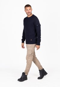Marineblauer gestrickter Pullover mit gerippten Bündchen und Saum, kombiniert mit beigen Cargo-Hosen. Schwarze Schnürstiefel. Modell läuft vor einem einfarbigen Hintergrund.