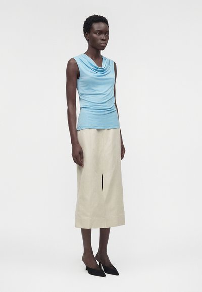 Γυναίκα με κοντά σγουρά μαλλιά που φοράει αμάνικο ανοιχτό μπλε τοπ με πέπλο, μπεζ midi φούστα με ραφή μπροστά και μαύρες μυτερές ψηλοτάκουνες γόβες.