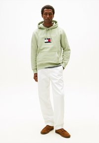 Felpa verde chiaro con logo, cordoncino e tasca frontale, abbinata a pantaloni bianchi e scarpe marroni; vestibilità comoda e materiale in cotone.