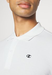 Hvit polo skjorte laget av bomull med to knapper og en svart brodert logo på venstre bryst. Glatt tekstur, brettet krage.