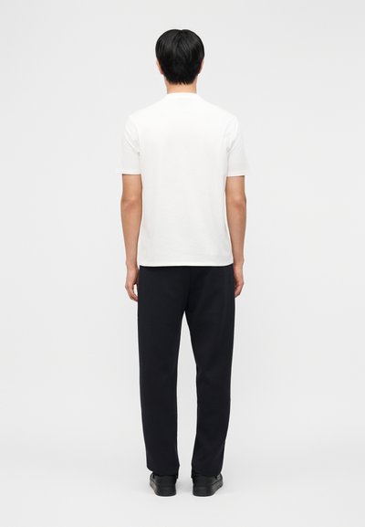 Άνδρας που στέκεται γυρισμένος με την πλάτη, φορώντας απλό λευκό κοντομάνικο πουκάμισο, μαύρο παντελόνι και μαύρα παπούτσια πάνω σε γκρι φόντο.