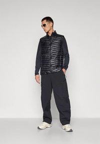 Crna prozračna jakna s teksturiranim završetkom, nošena preko crne majice dugih rukava i uparena s širokim crnim hlačama i tenisicama.