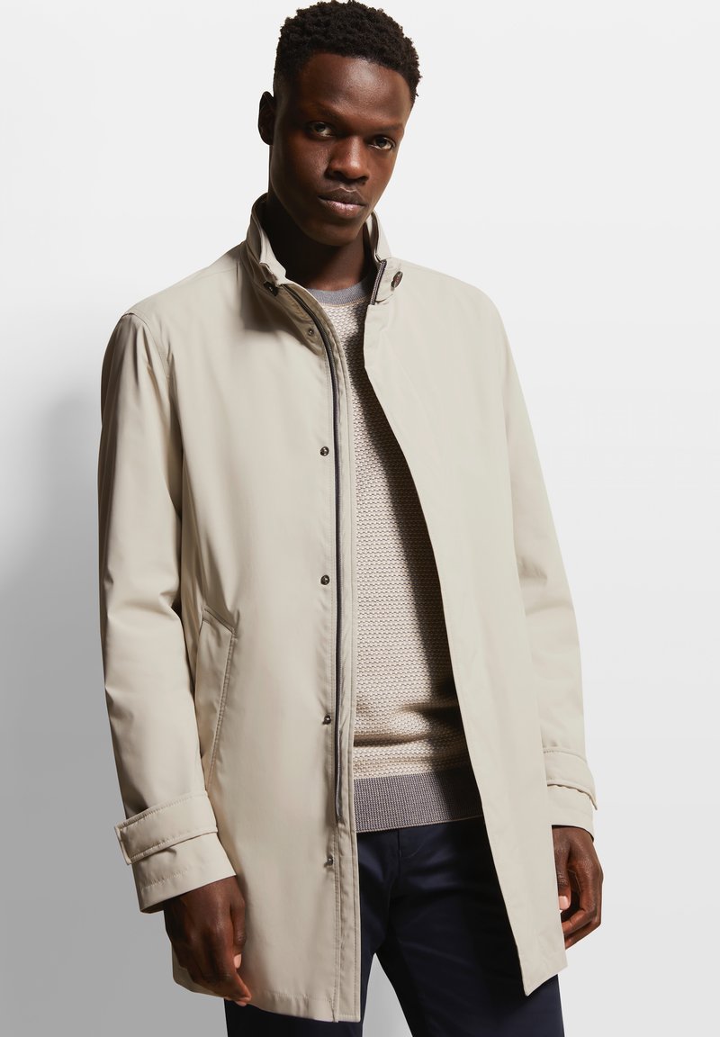 Manteau beige clair avec un col montant, fermeture à pression, poches latérales et texture lisse. Porté sur un pull en laine gris texturé.