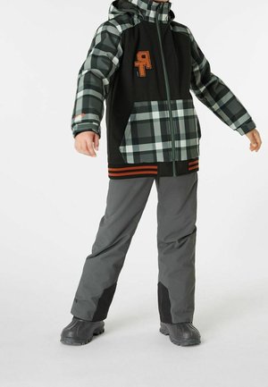 Chaqueta de cuadros en negro y verde con un logo naranja, combinada con pantalones grises y detalles en negro; material impermeable, capucha ajustable y bolsillos.