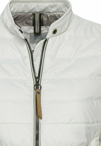 Veste matelassée blanche avec col montant, fermeture éclair noire avec tirette marron et pression au niveau du cou, montrant l’étiquette camel active à l’intérieur.