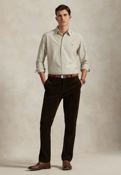 Tânăr stând cu mâinile în buzunare, purtând o cămașă bej în carouri, pantaloni maro închis, curea maro, ceas și pantofi maro pe un fundal simplu.