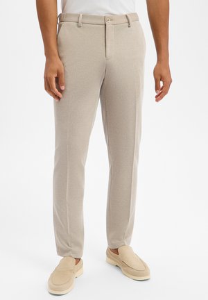 Beige, slim-fit Hose mit Gürtelschlaufen und Knopf, getragen von einer Person mit beigen Slip-on Schuhen und weißem Top vor weißem Hintergrund.