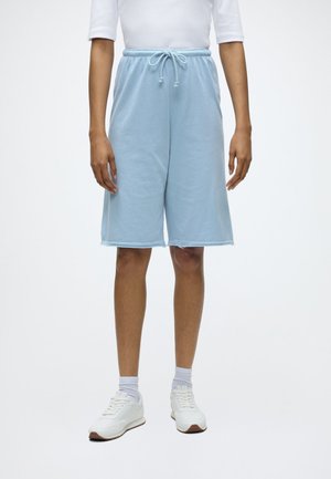 Persoană purtând pantaloni scurți bleu deschis până la genunchi cu șnur, șosete albe, adidași albi și un pulover alb cu mânecă lungă pe un fundal simplu.