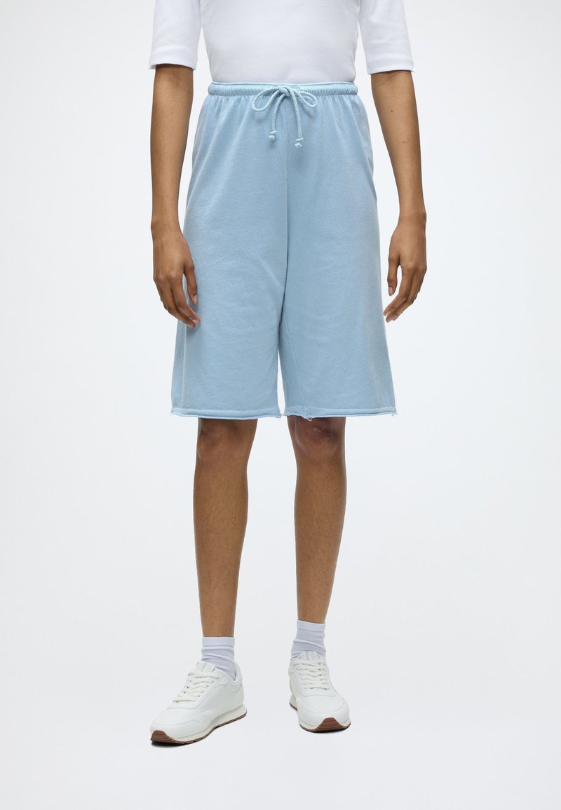 Άτομο που φοράει φωτεινά μπλε σορτς μέχρι το γόνατο με κορδόνι, λευκές κάλτσες, λευκά αθλητικά παπούτσια και λευκό μακρυμάνικο μπλουζάκι σε μονόχρωμο φόντο.