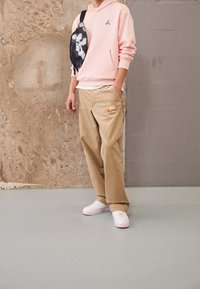 Felpa con cappuccio rosa chiaro con logo, pantaloni beige con un logo arancione, sneakers bianche e una borsa a tracolla nera in camouflage.