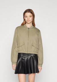 Olivovo zelená bomber bunda so zipsom vpredu, elastickými manžetami a dvoma bočnými vreckami. Nosí sa cez čiernu plisovanú sukňu z imitácie kože.