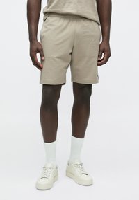 Bēša kokvilnas šorti ar elastīgu jostasvietu, sānu kabatām un ērtu piegriezumu, kopā ar baltām zeķēm un krēmkrāsas sporta apaviem.