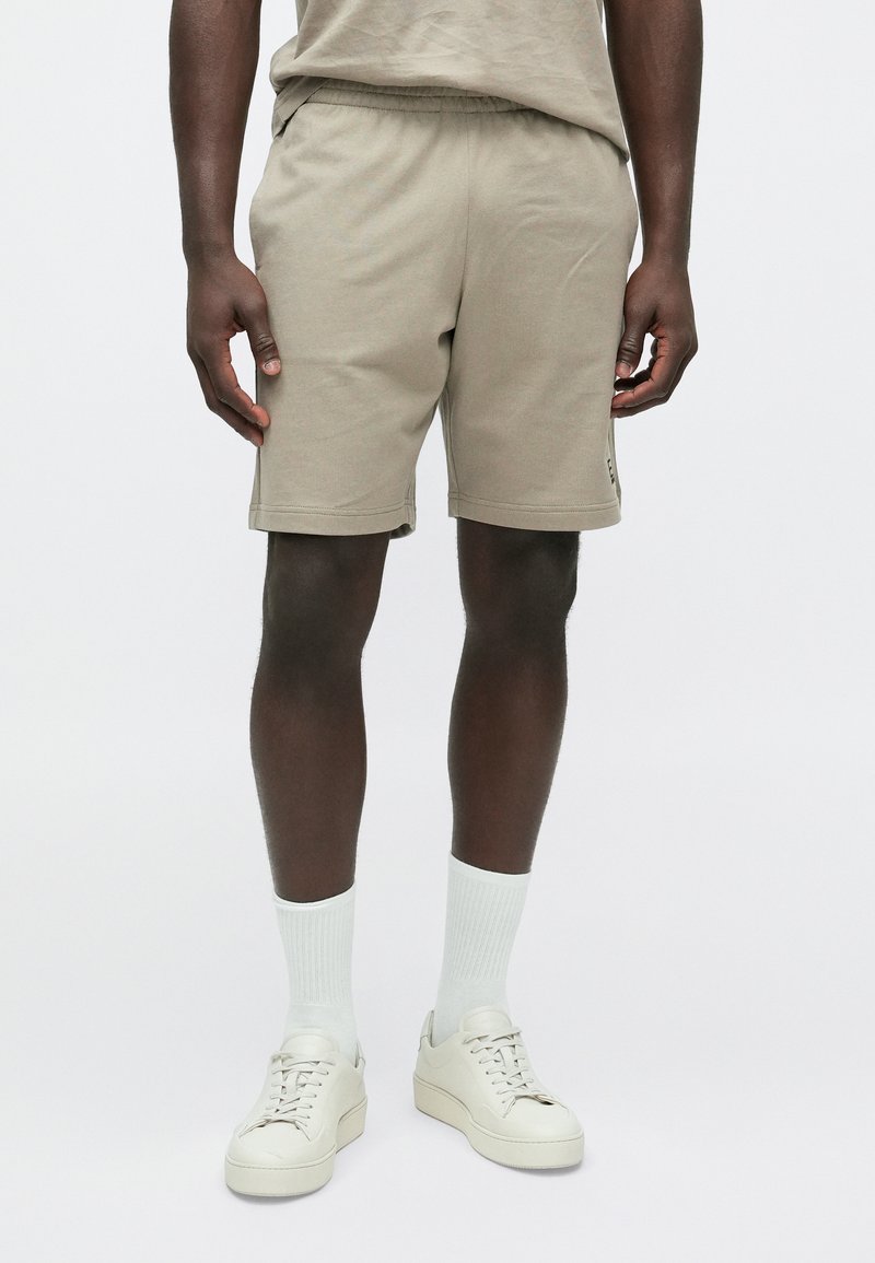 Bēša kokvilnas šorti ar elastīgu jostasvietu, sānu kabatām un ērtu piegriezumu, kopā ar baltām zeķēm un krēmkrāsas sporta apaviem.