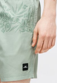 Ανοιχτό πράσινο σορτς με διακριτικό λουλουδάτο μοτίβο και ταμπέλα με το λογότυπο O'Neill, εμφανίζεται δίπλα σε ένα χαλαρό χέρι στο ύψος του μηρού.