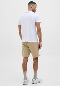 T-shirt blanche à manches courtes, shorts beiges, chaussettes grises et baskets blanches. Design droit, texture lisse, style minimaliste.