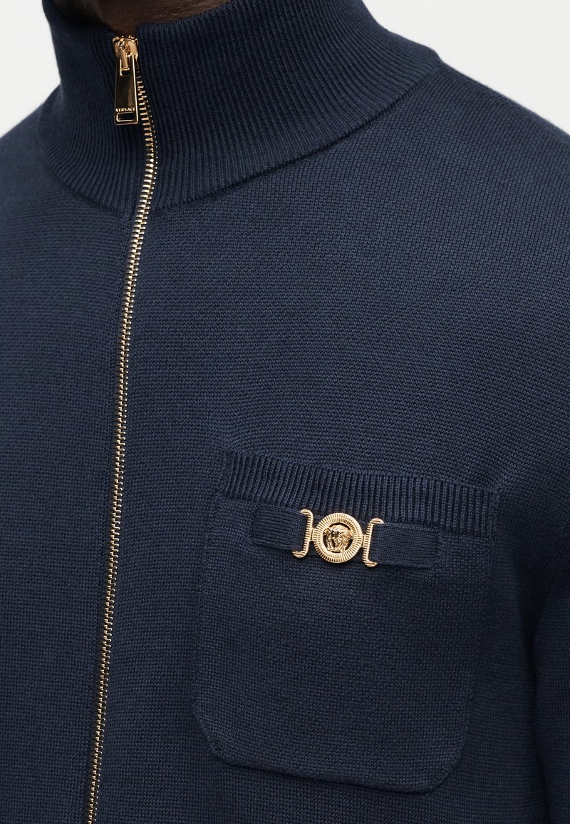 Pull en maille marine à fermeture éclair avec col montant, fermeture éclair dorée, petite poche poitrine et emblème décoratif doré de Méduse sur la poche.