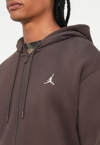 Osoba v hned ej zipovej mikine s kapucňou a bielym logom Jumpman na hrudi, s vrkočmi a camouflage tričkom pod ňou.