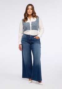 Camicia bianca con bottoni e pannello testurizzato blu, abbinata a jeans di denim blu a gamba larga. Il modello sta in piedi con sicurezza con una mano nella tasca.