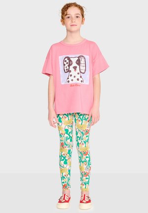 Girl with curly red hair wearing a pink dog-illustrated t-shirt, colorful floral leggings, and red slip-on shoes, standing against a plain background.