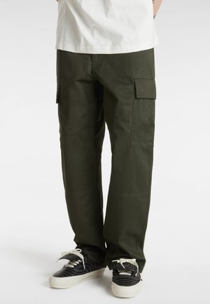 Personne portant un pantalon cargo vert foncé avec des poches latérales et des baskets noir et blanc, debout devant un fond clair uni.