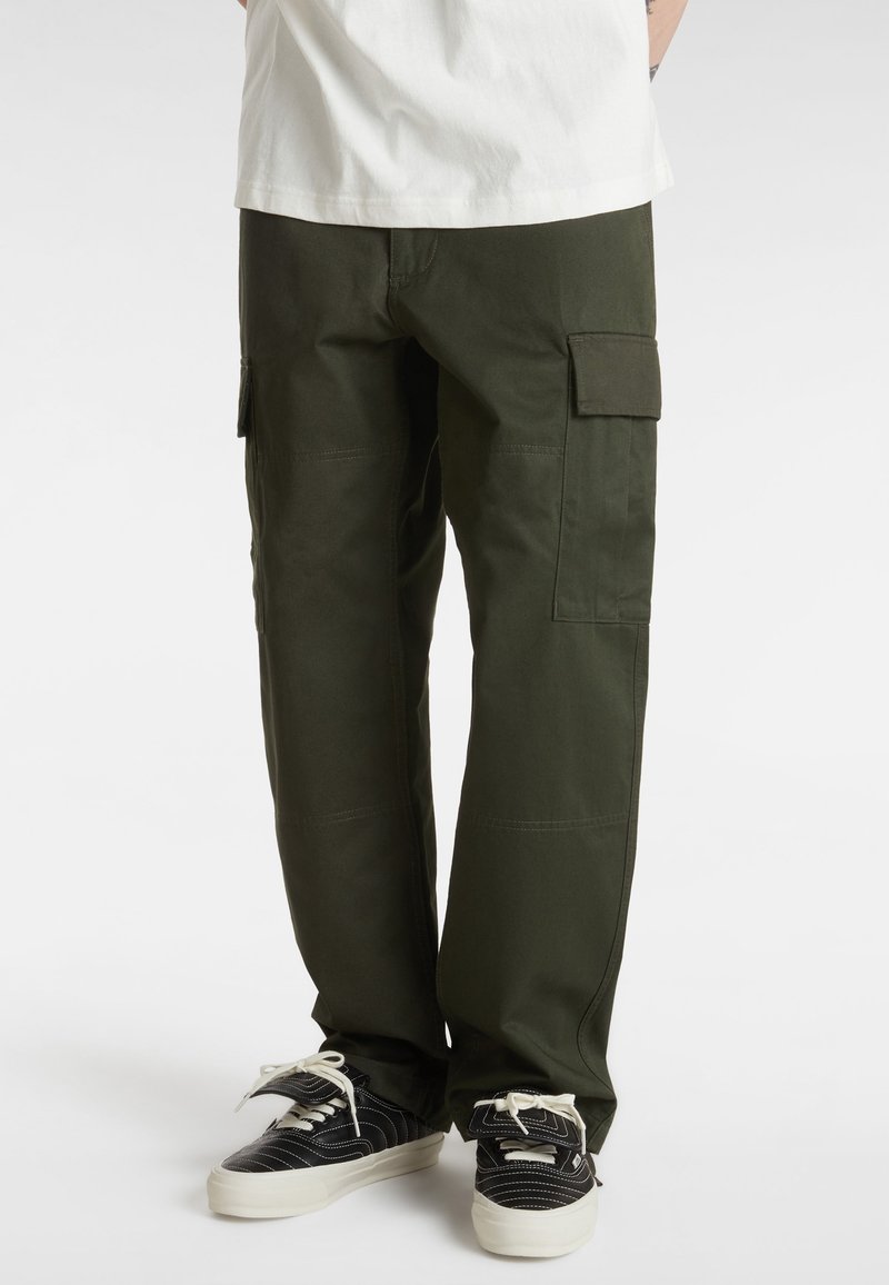 Personne portant un pantalon cargo vert foncé avec des poches latérales et des baskets noir et blanc, debout devant un fond clair uni.
