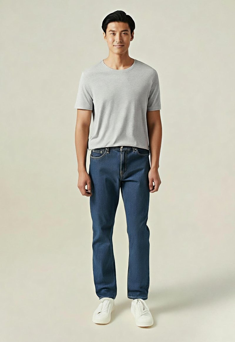 Hombre de pie, vestido con una camiseta de manga corta gris clara, jeans azules y zapatillas blancas, frente a un fondo ligero y sencillo.