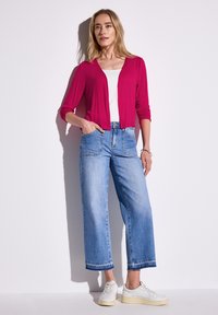Un cardigan léger rose sur un haut blanc, associé à un jean large bleu délavé. Des baskets blanches complètent la tenue.